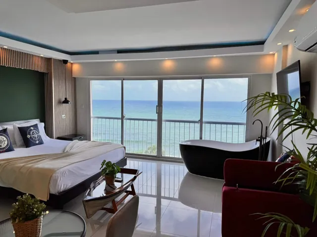 Studio with a black freestanding bathtub, king bed and a wall of windows on the sea
