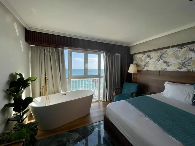 Studio with a freestanding bathtub, a plant and a teal-trimmed bed facing the sea