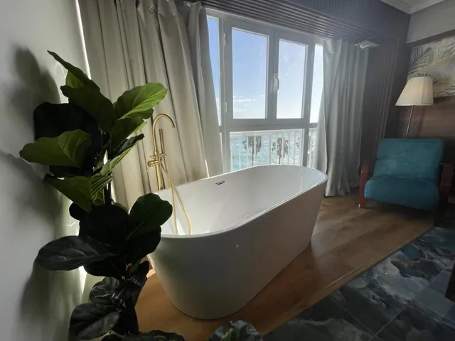 Bathtub with a brass faucet beside a fiddle-leaf fig and the sea view