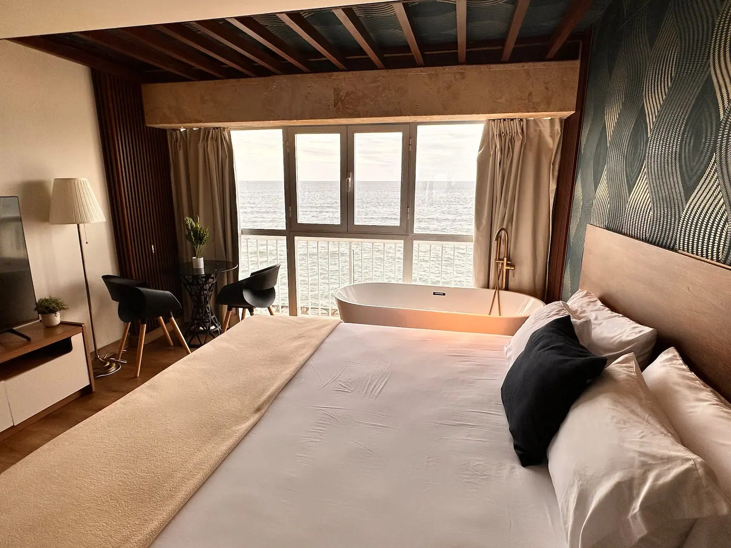 Studio with wood beams, a large bed and a tub at the window